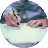 Eine Hand mit einem Stift schreibt auf ein Blatt Papier. Die Person trägt ein hellblaues Hemd.