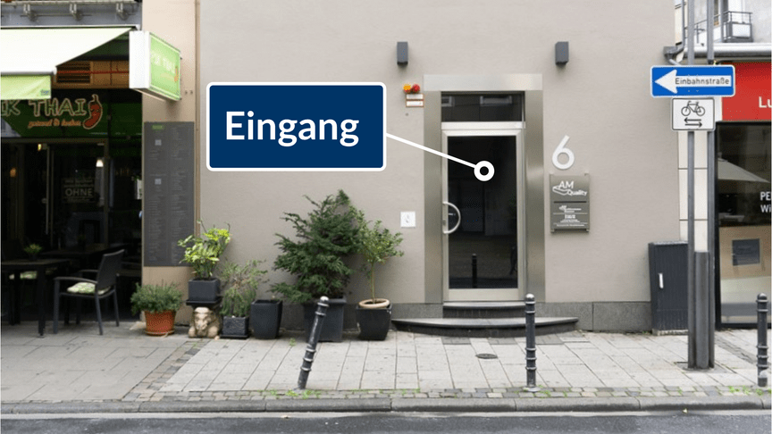 Eingang zu einem Geschäft mit gläserner Tür und Beschilderung, umgeben von Pflanzen und Straßenschildern.