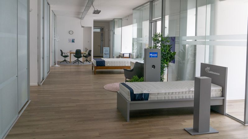 Zwei moderne Betten mit Matratzen in einem hellen Showroom, umgeben von Pflanzen und Sitzgelegenheiten.