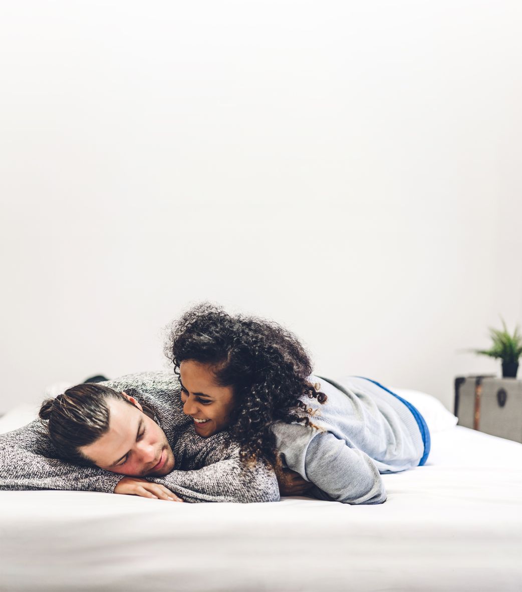 Ein Paar liegt entspannt auf einem Bett, lächelt sich an und genießt die Nähe. Die Frau hat lockiges Haar und trägt einen grauen Pullover.