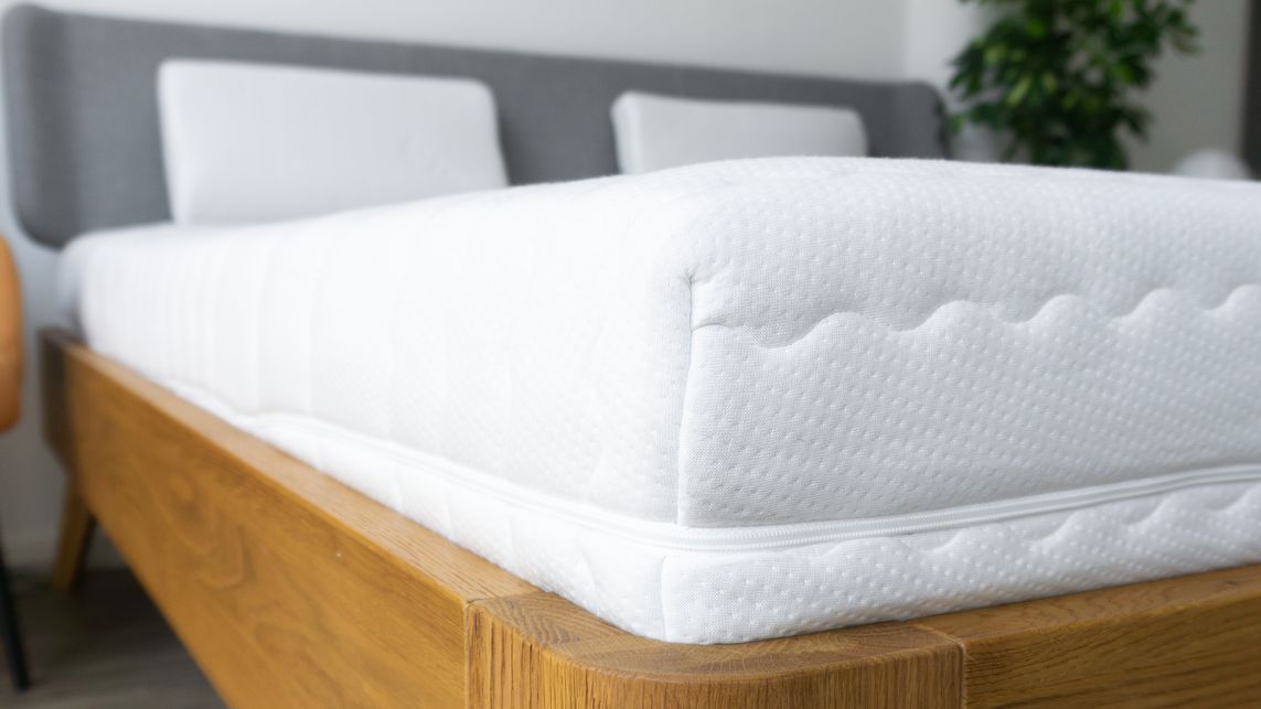 Eine weiße Matratze mit gestepptem Bezug auf einem Holzbettrahmen, seitliche Ansicht.