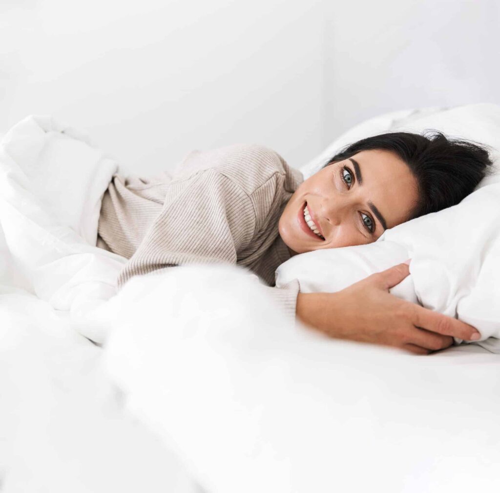Eine lächelnde Frau in einem beigen Pullover liegt entspannt auf weißen Kissen und Decken.