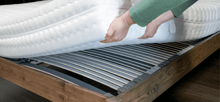 Eine Person hebt eine Matratze an, um einen Lattenrost aus Holz mit flexiblen Leisten zu zeigen.