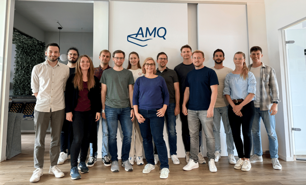 Teamfoto von AMQ, das aus 14 Personen besteht, die in lässiger Kleidung vor einem Logo stehen.