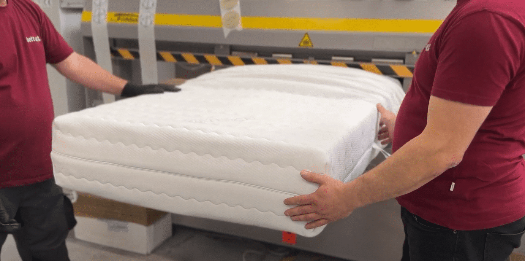 Zwei Personen heben eine weiße Matratze mit gestepptem Bezug, bereit für den Transport.