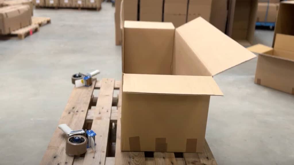Ein offener, brauner Karton auf einer Holzpalette, daneben zwei Klebebandabroller.