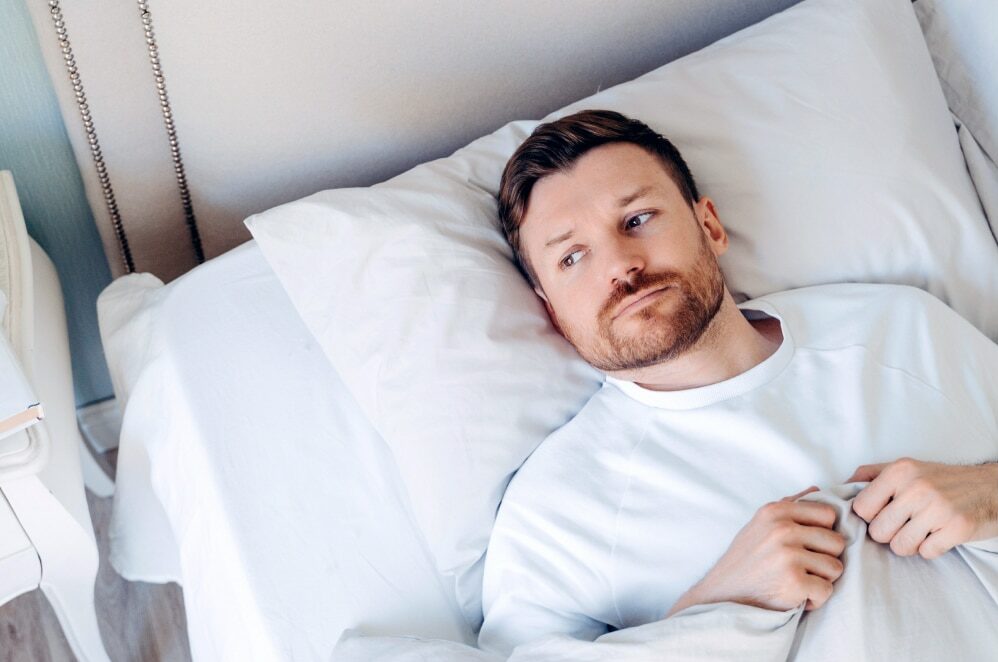 Ein Mann mit Bart liegt in einem Bett und schaut nachdenklich zur Seite, bekleidet mit einem weißen Oberteil.