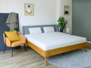 Ein modernes Schlafzimmer mit einem Holzbett, grauem Kopfteil, zwei weißen Kissen und einem beigen Sessel mit gelbem Kissen.