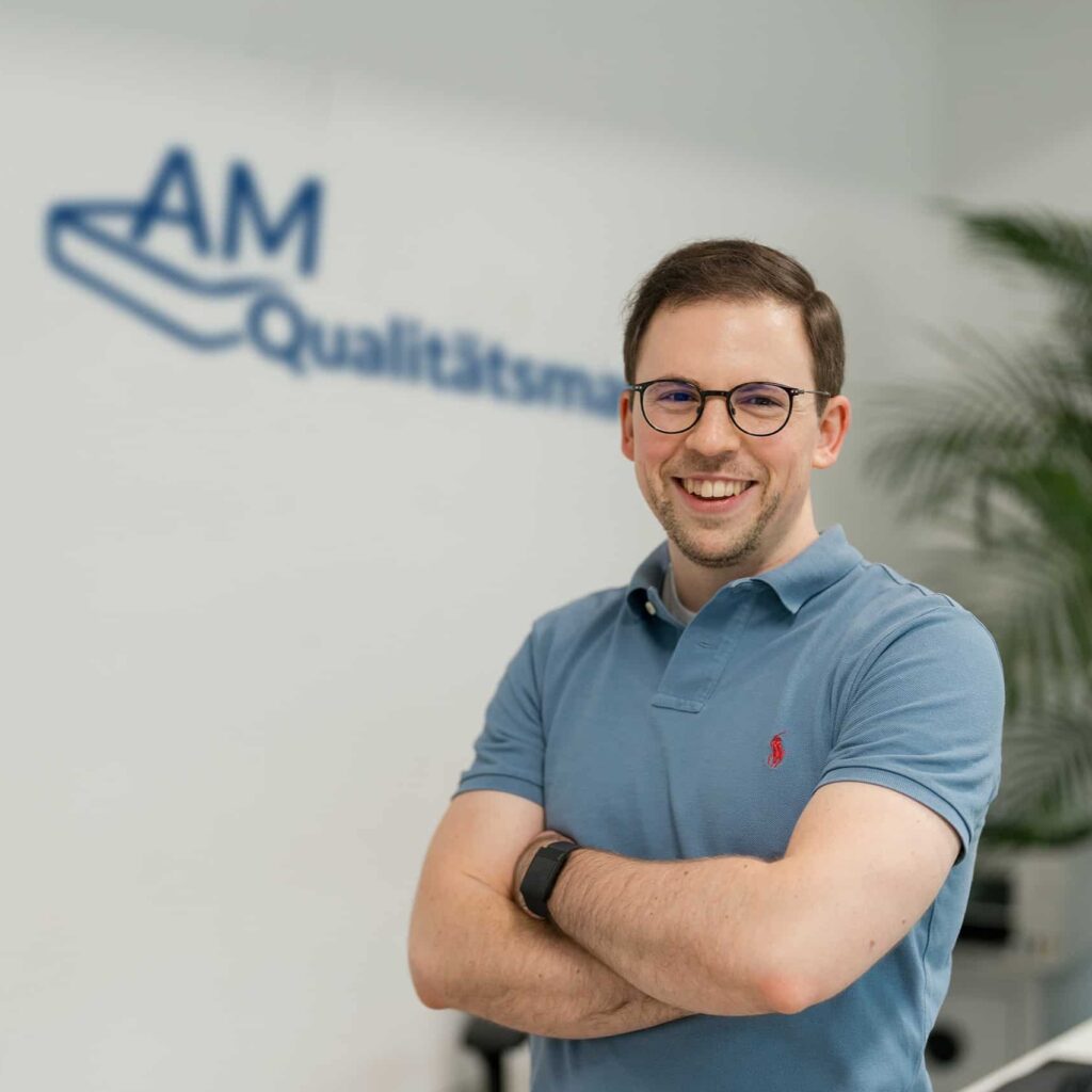 Ein lächelnder Mann mit Brille trägt ein hellblaues Polo-Shirt und steht mit verschränkten Armen vor einer Wand mit dem Schriftzug "AM Qualitätsmanagement".
