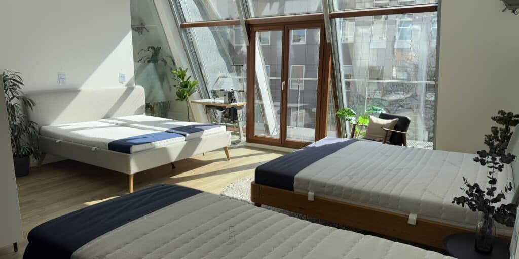 Drei Betten mit Matratzen im Showroom in Köln von AM Qualitätsmatratzen, ein helles Bett mit grauem Bezug und zwei Betten mit dunkelblauen Akzenten.