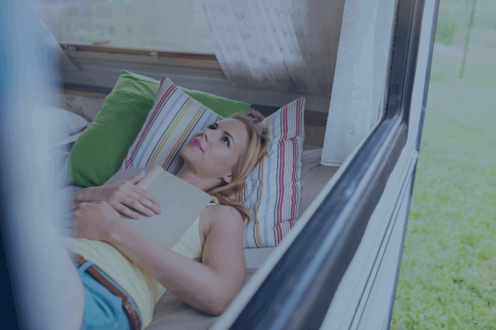 Junge Frau mit geschlossenen Augen liegt entspannt auf einem Fensterbank-Kissen, umgeben von bunten Kissen.