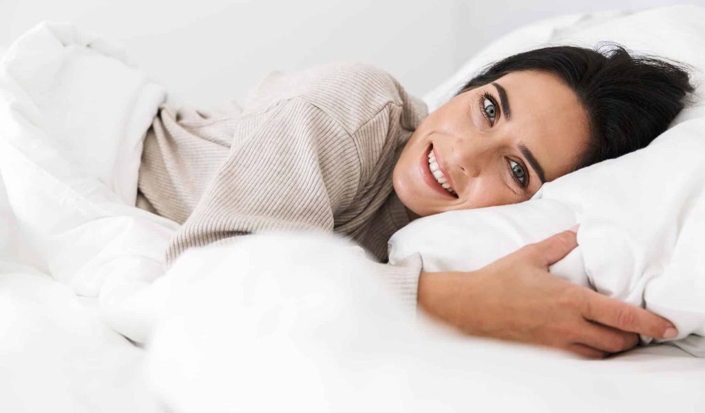 Eine lächelnde Frau in einem beigen Pullover liegt entspannt auf weißen Kissen und Decken.