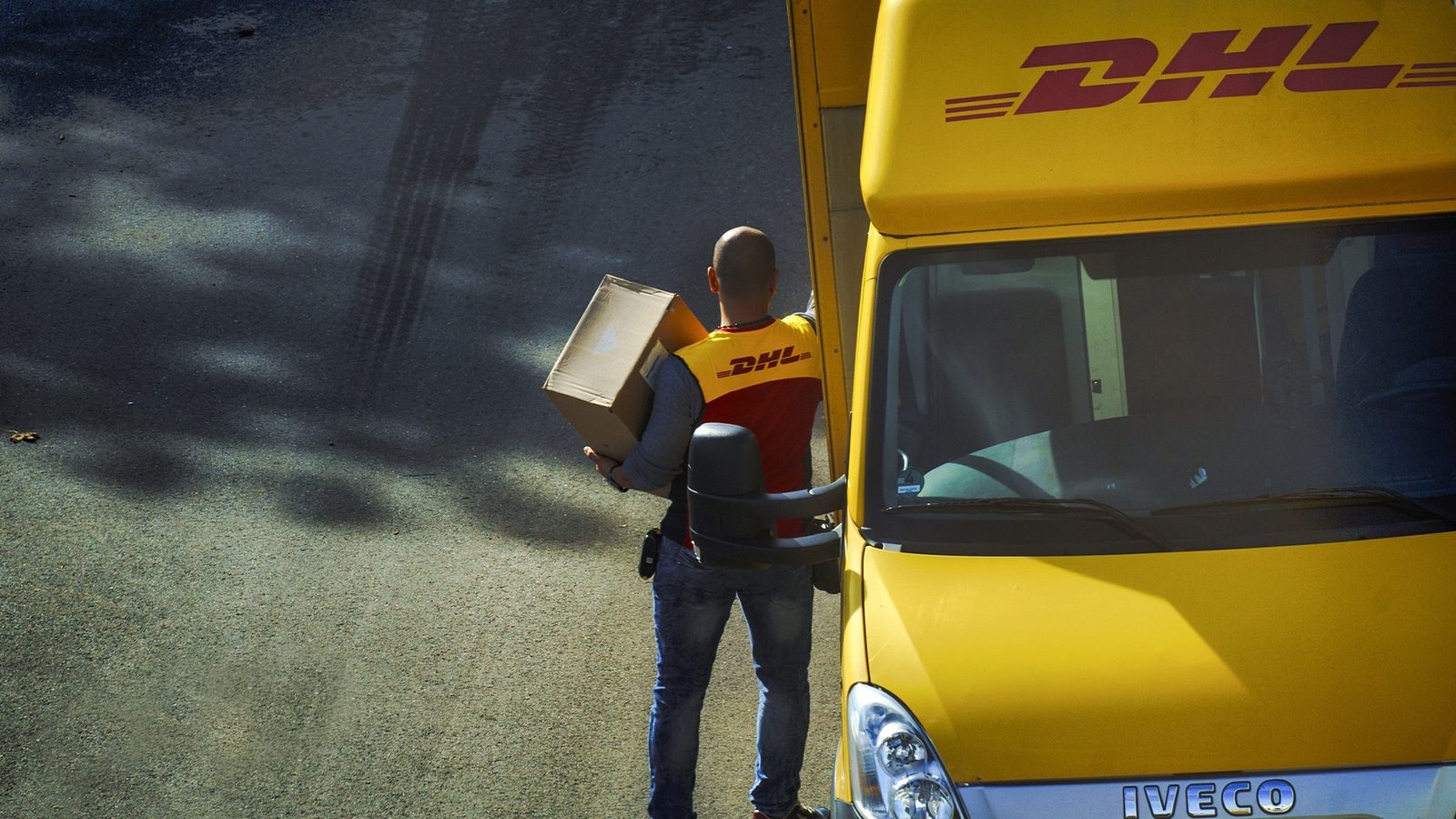 Ein DHL-Zusteller trägt ein Paket und steht neben einem gelben Lieferwagen.